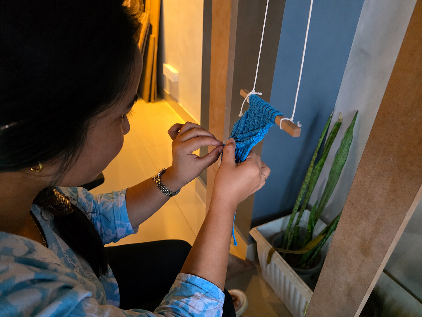 Artisan woman hand-knotting a blue macramé piece on a wooden dowel.